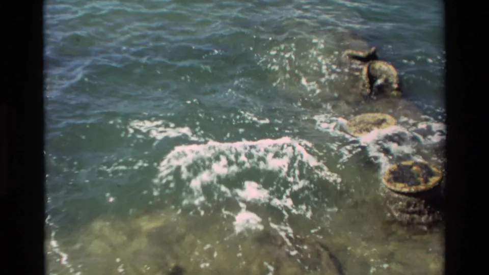 1981 Hawaii 8mm frame looking down at clear water, white foam, and rocks or mooring posts beside the waterfront structure.