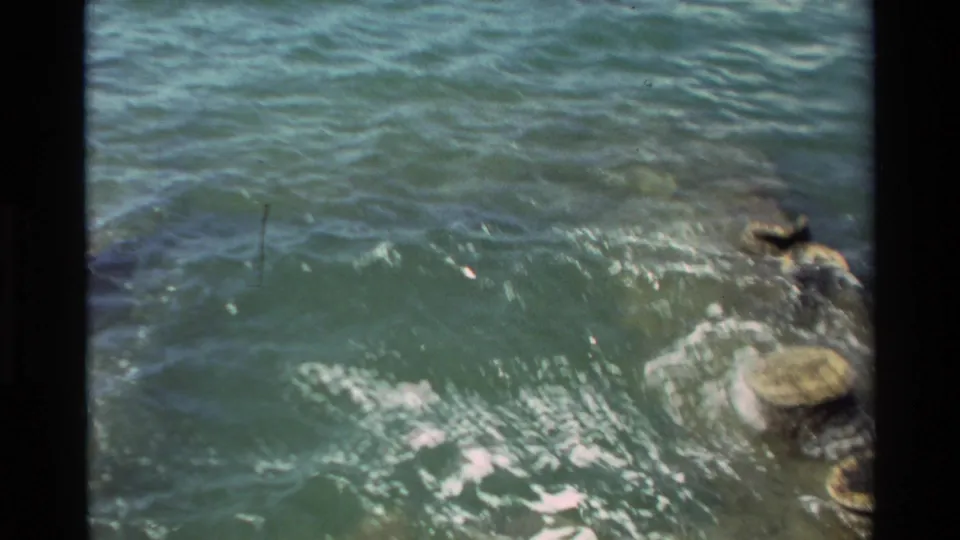 1981 Hawaii 8mm frame showing waves moving around dark shoreline rocks, with white foam and soft home-movie grain.
