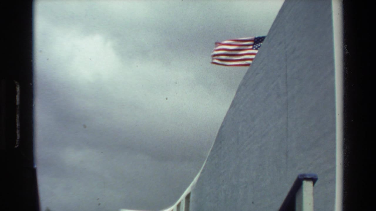 American flag flying beside a white curved memorial wall under cloudy sky.