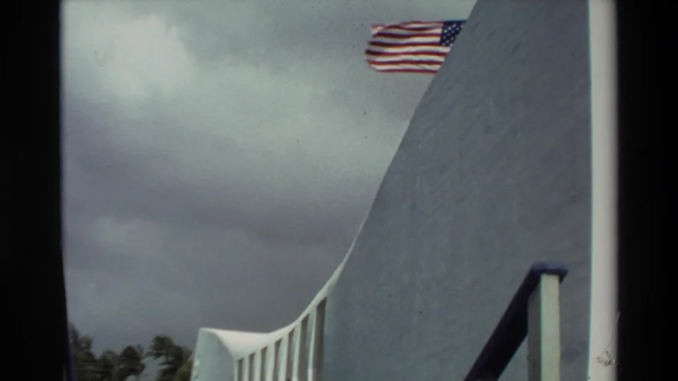 1981 Hawaii 8mm frame of the USS Arizona Memorial wall, flag, railing, cloudy sky, and distant palm trees.