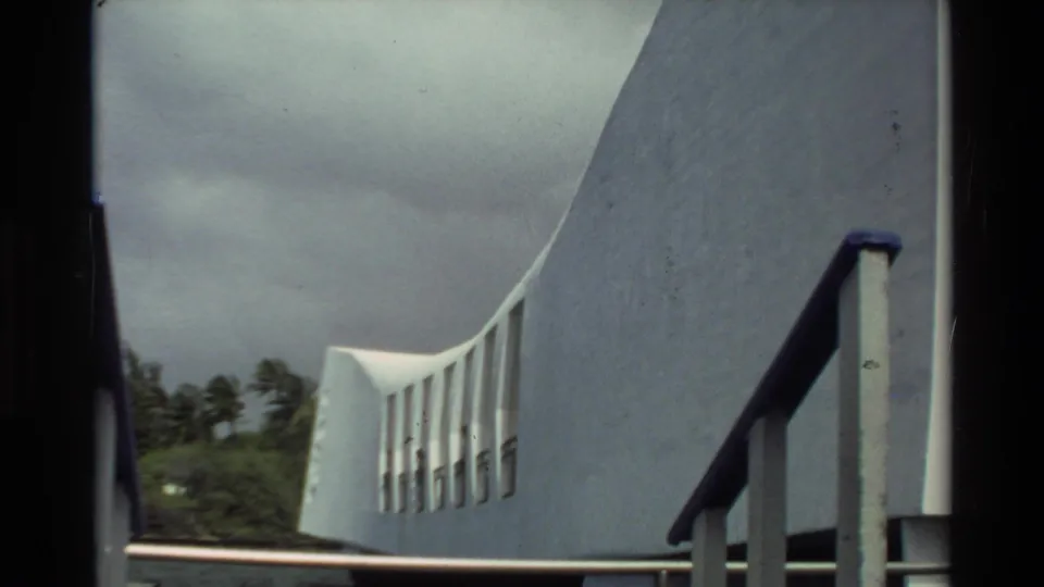 1981 Hawaii frame showing the USS Arizona Memorial exterior wall and railings with distant palms under overcast sky.