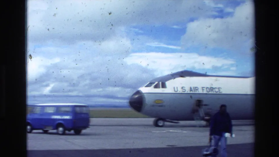 1981 New Zealand airport tarmac scene with a U.S. Air Force transport aircraft, a blue van, and civilians walking nearby.