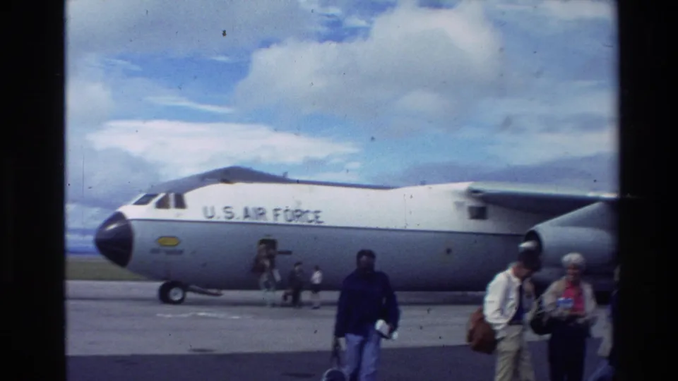 1981 New Zealand home-movie frame showing civilians passing near a parked U.S. Air Force transport aircraft under a cloudy sky.