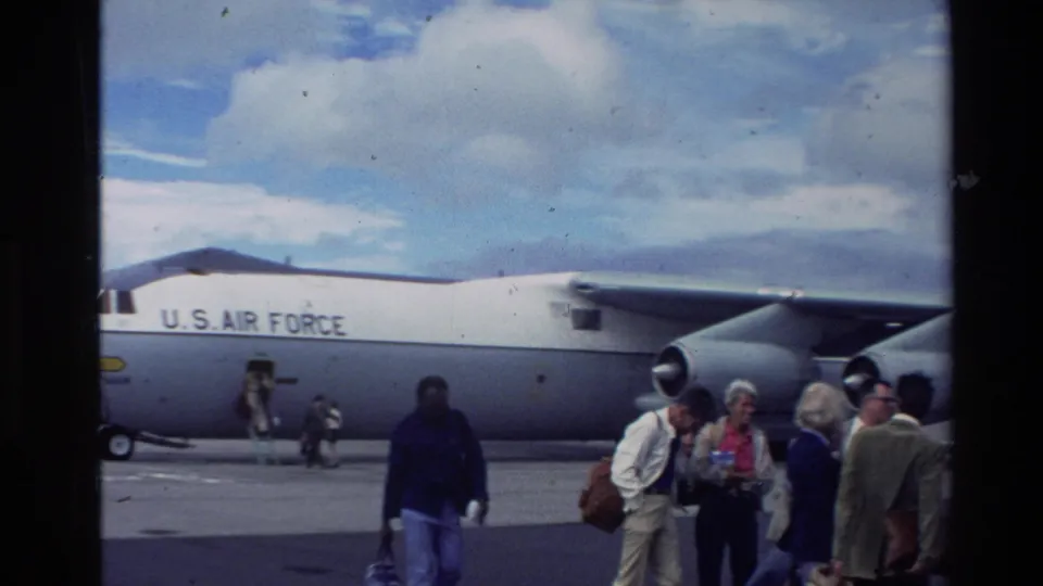 1981 New Zealand home-movie frame showing adults with bags beside a large gray U.S. Air Force transport aircraft on an airfield.