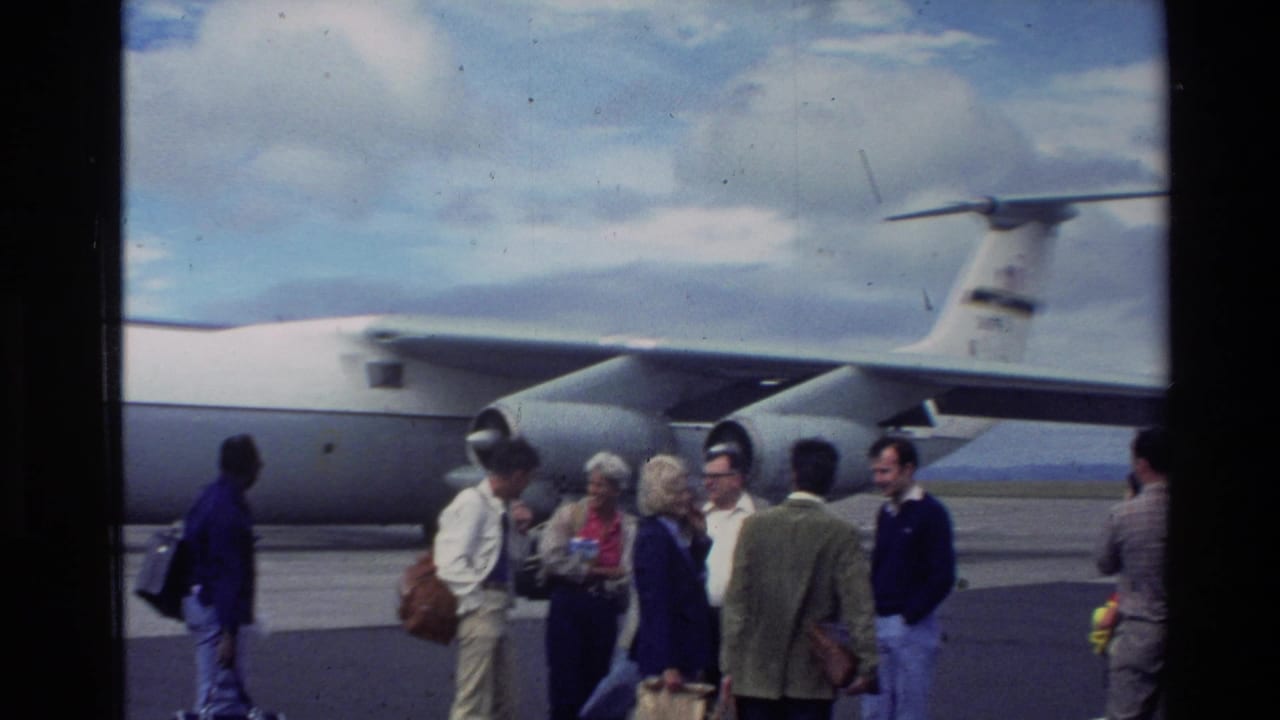 Adults in 1980s casual clothing converse on the tarmac beside a gray U.S. Air Force transport aircraft in New Zealand.
