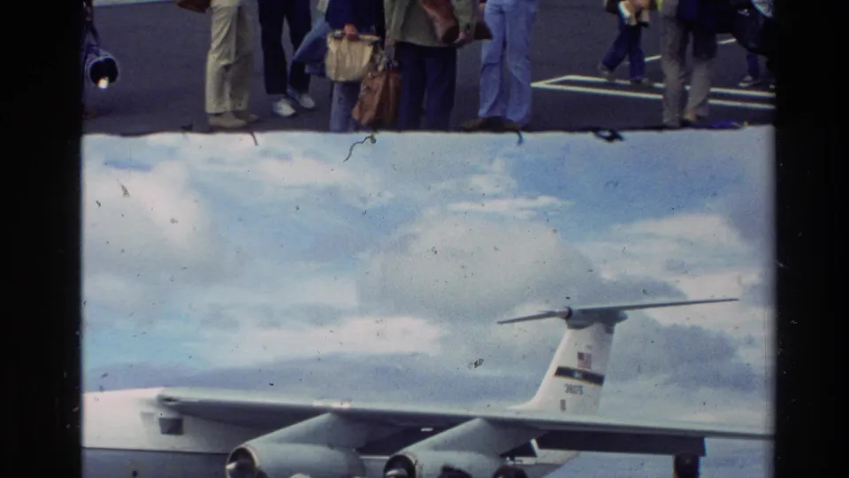 Home-movie view of a large gray U.S. Air Force transport aircraft on a New Zealand airfield, with travelers partly visible in front.