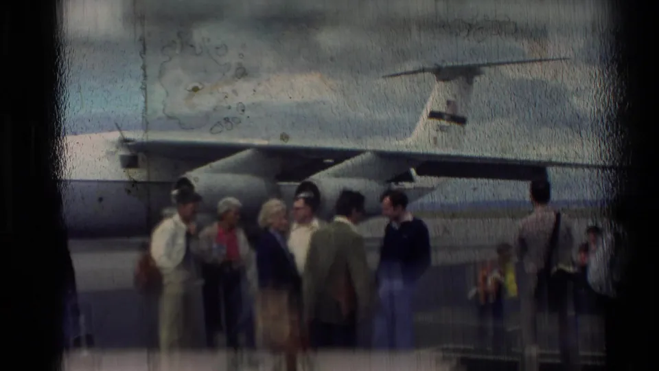 Soft 8mm frame from 1981 New Zealand showing adults gathered near a gray transport aircraft, with the film surface visibly worn.