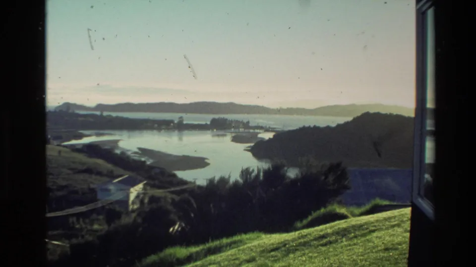 1981 New Zealand 8mm view of a calm coastal inlet, low hills, grassy foreground, and distant houses framed by a dark window edge.