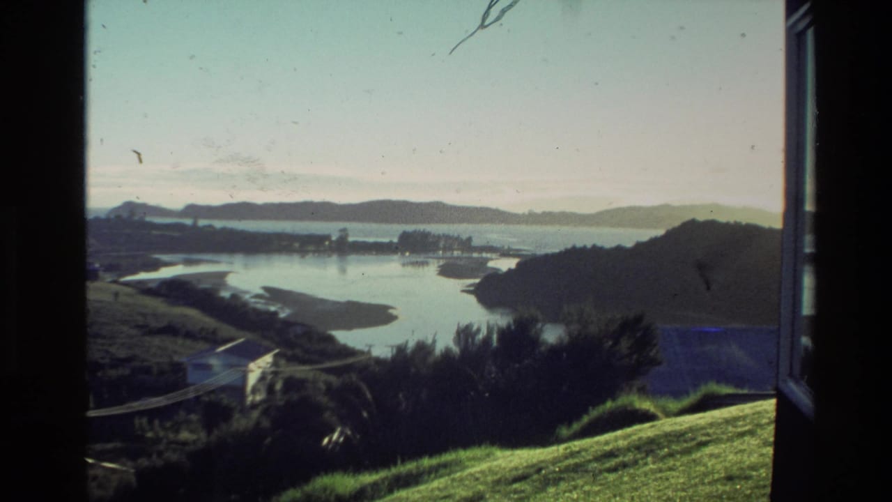 A similar 1981 New Zealand home-movie view shows still water, low hills, small buildings, and green foreground through a window frame.