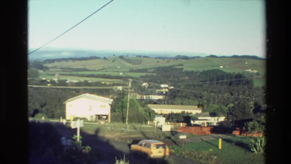 1981 New Zealand rural settlement seen from above, with modest buildings, power lines, green hills, and a parked yellow car.
