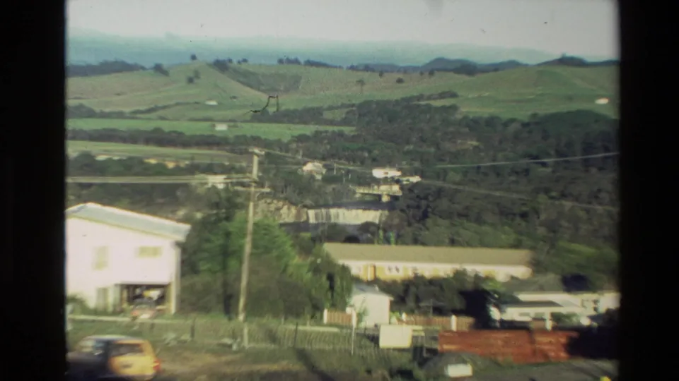 Soft 8mm frame of a New Zealand rural hillside settlement, showing buildings, utility lines, fields, trees, and a yellow car.