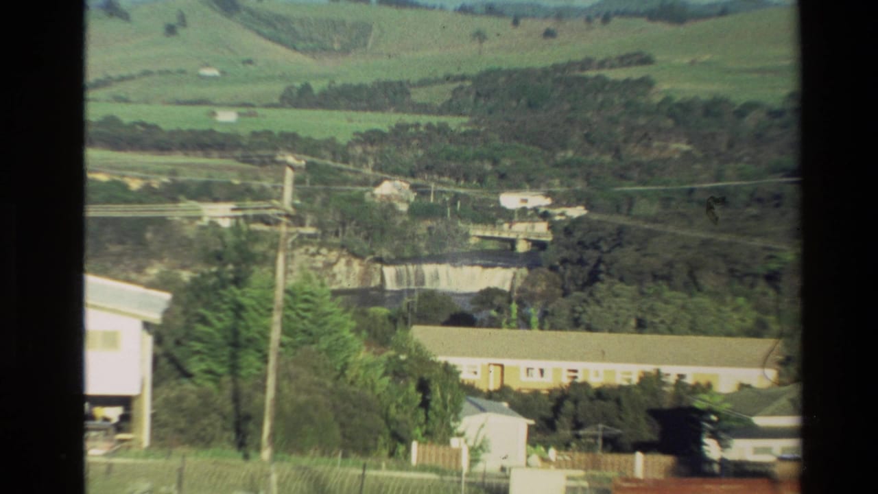 Rural hillside view with buildings, power poles, river, bridge, and waterfall in the distance.