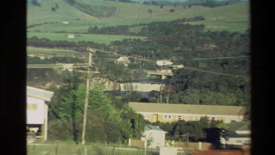 1981 New Zealand 8mm frame showing a rural hillside overlook with buildings, power lines, a bridge, and a distant waterfall.