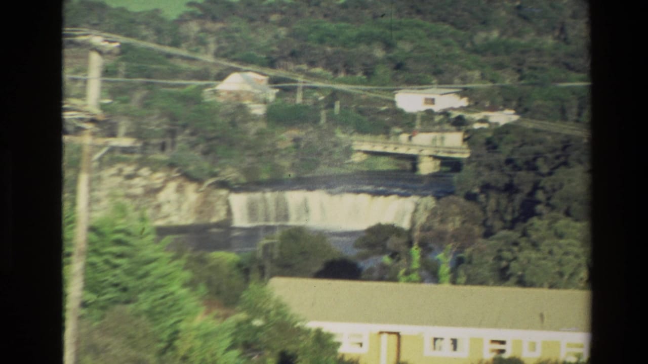 1981 New Zealand 8mm frame closer on the river crossing, showing a bridge, waterfall, trees, power lines, and nearby buildings.