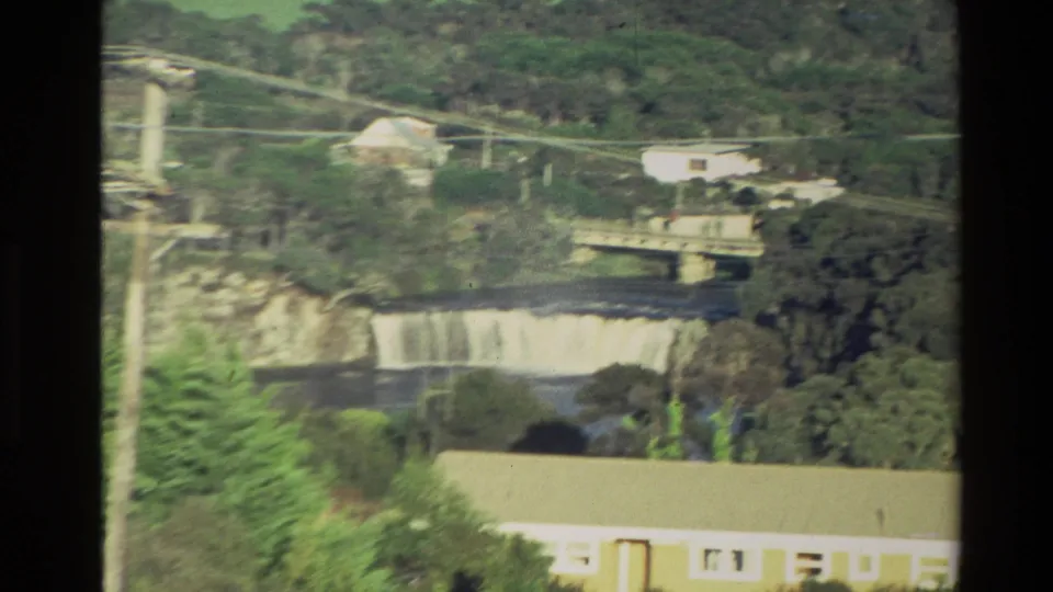 1981 New Zealand 8mm frame closer on the river crossing, showing a bridge, waterfall, trees, power lines, and nearby buildings.