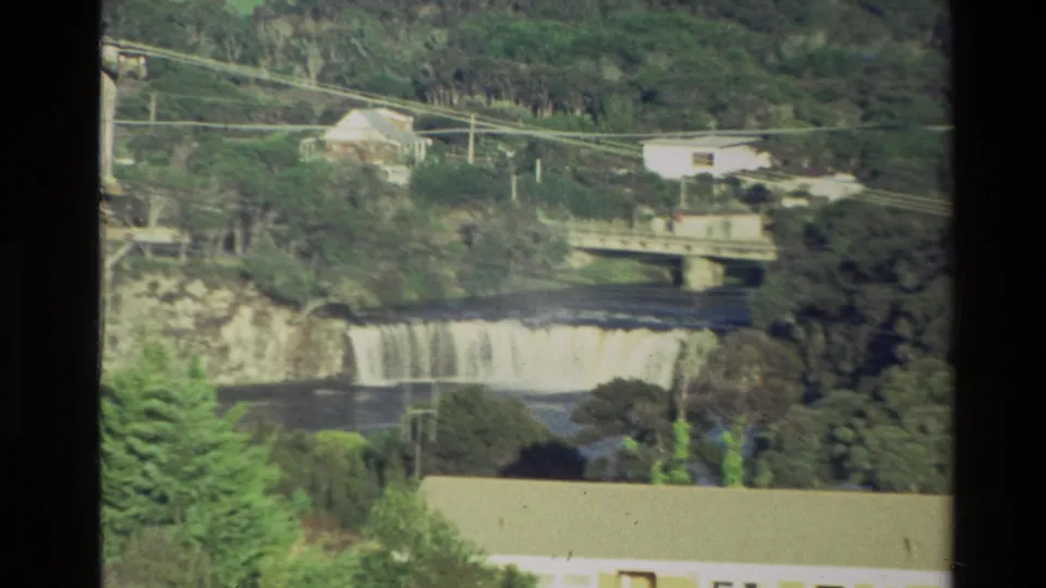 1981 New Zealand 8mm frame showing the zoom continuing toward a waterfall below a bridge, with trees, power lines, and low buildings.