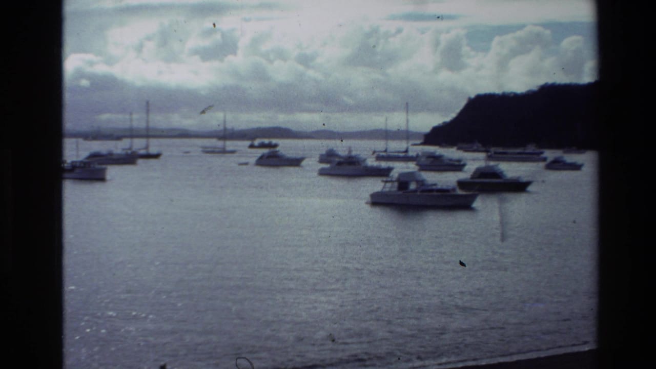 1981 New Zealand coastal home-movie frame with anchored boats, sailboat masts, calm water, and a dark hill at the bay edge.