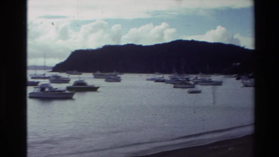 1981 New Zealand home-movie frame with a wider view of many boats at anchor across a calm bay under clouds.
