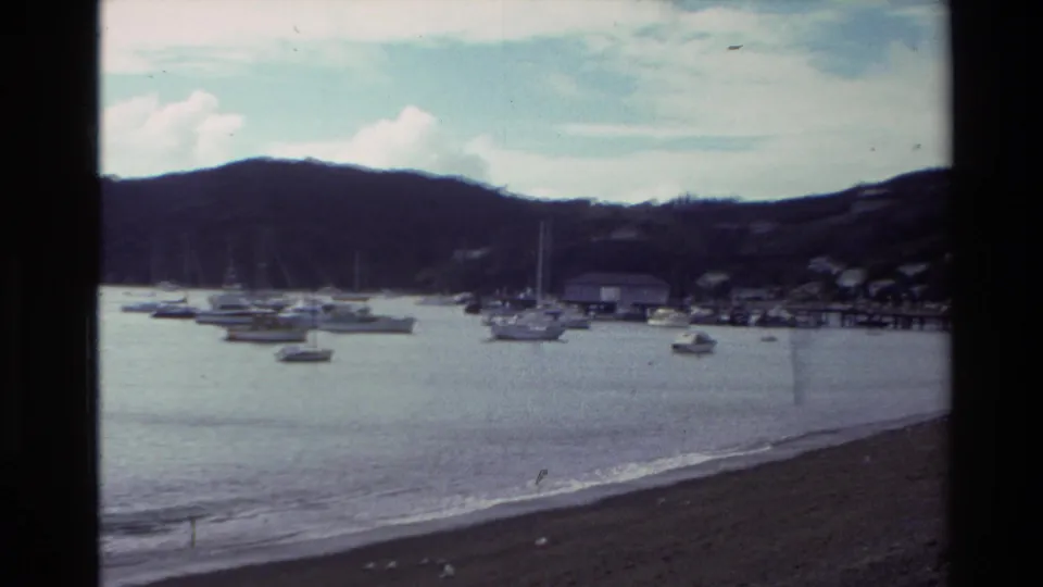 1981 New Zealand coastal bay view with anchored sailboats, a curved beach, low waves, hills, and distant shoreline structures.