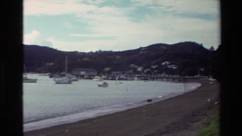 1981 New Zealand 8mm frame showing a quiet beach, calm bay water, anchored boats, and tree-covered hills in the distance.