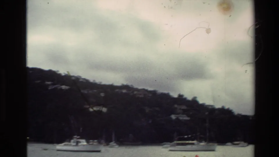 1981 New Zealand home-movie frame showing anchored boats on calm water beneath a tree-covered hillside and overcast sky.