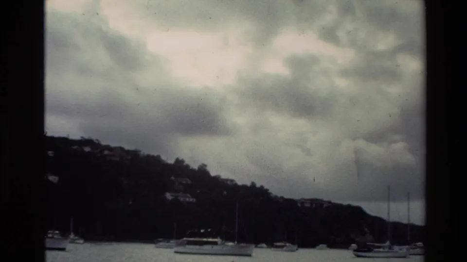 1981 New Zealand harbor view with moored boats, shoreline hillside, scattered houses, and heavy clouds in an 8mm frame.