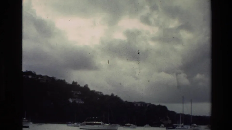 1981 New Zealand 8mm frame of a quiet harbor, with boats at anchor below a wooded hillside and dramatic cloud cover.