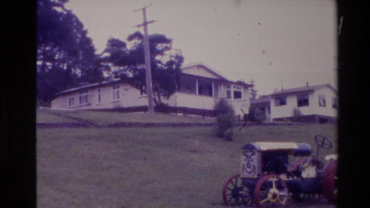 1981 New Zealand home-movie frame showing a rural house on a grassy slope, utility poles, trees, and an older decorative tractor-like vehicle.