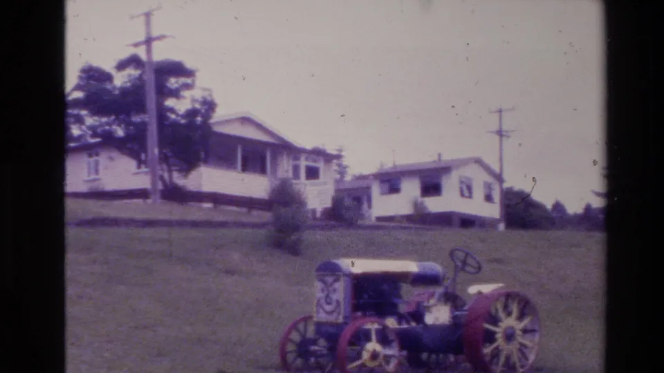 1981 New Zealand frame of a hilltop rural house, roadside-style utility poles, trees, and an older tractor-like vehicle partly visible in front.