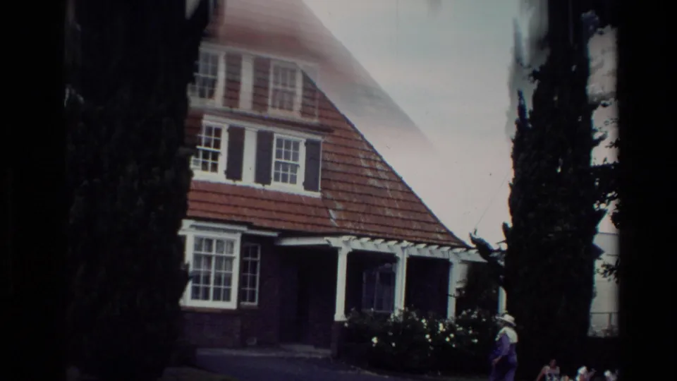 1981 New Zealand home-movie view of a brick house with red tiled roof, white trim, tall evergreens, garden, and a person nearby.