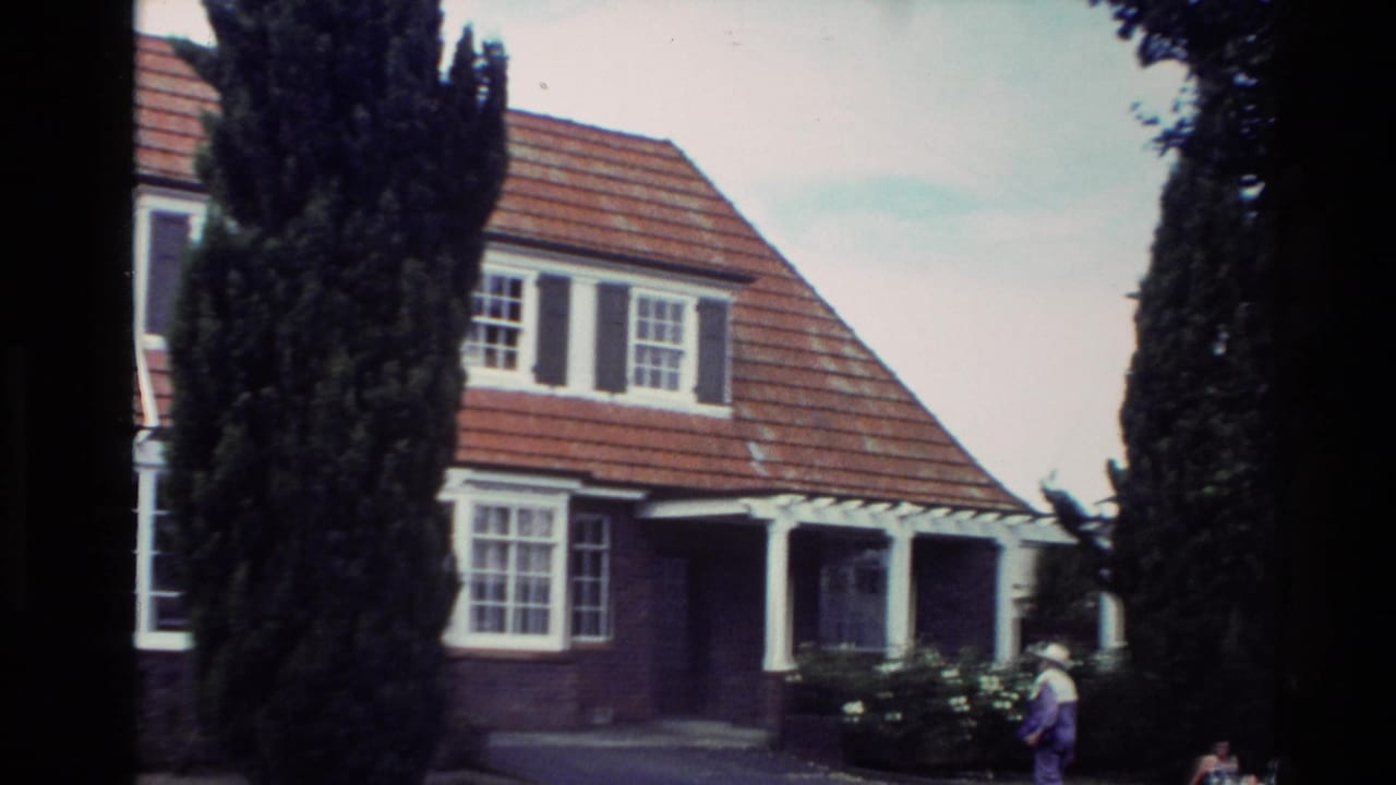 Brick bungalow with red tiled roof, white windows, cypress trees, and a person near the front.