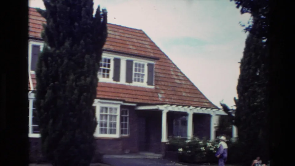 1981 New Zealand home-movie frame of a brick bungalow frontage with red tiled roof, white trim, tall cypress trees, and a person outside.