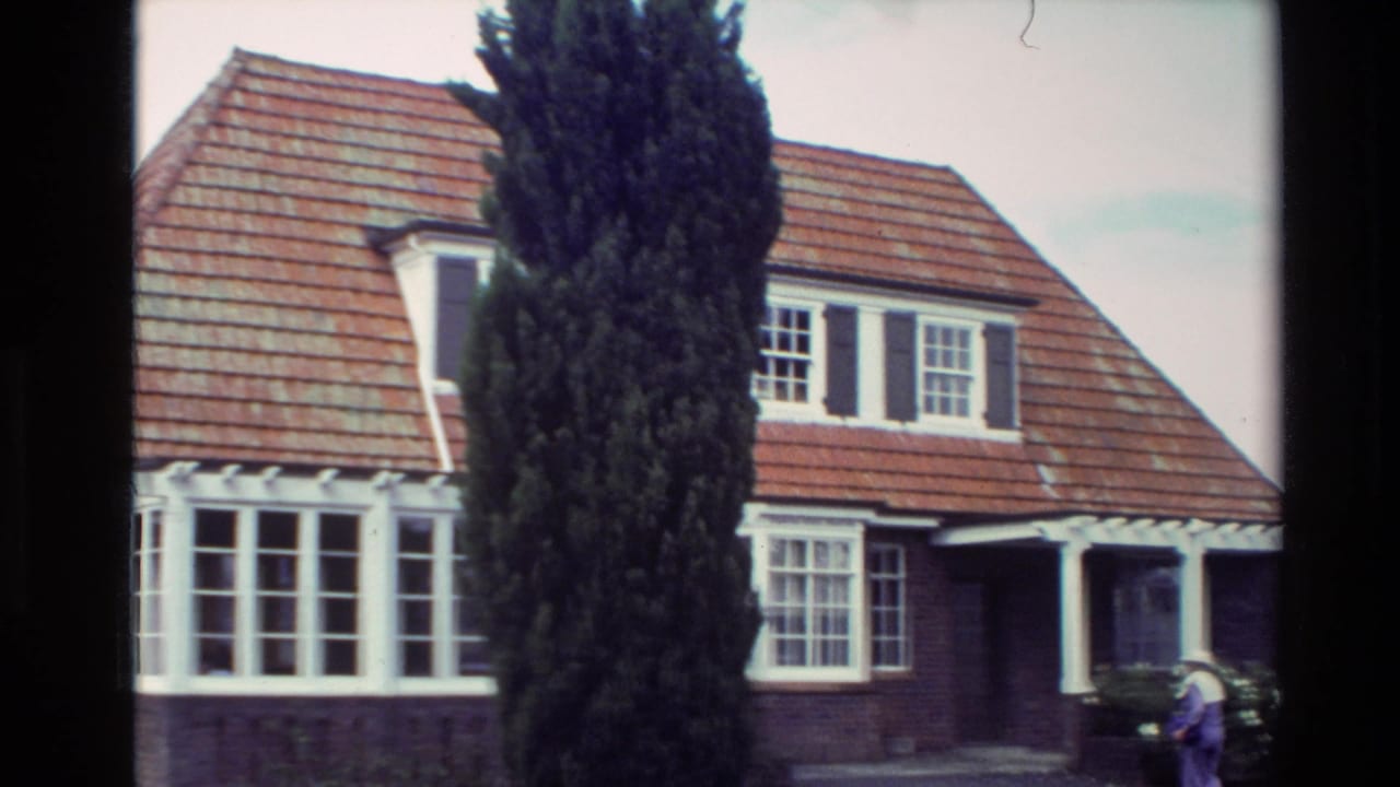 Closer 8mm view of the New Zealand bungalow, showing red roof tiles, dormer windows, white trim, a cypress tree, and a person outside.