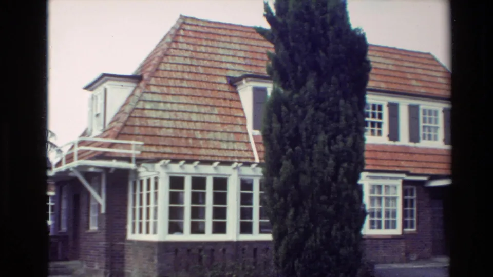 1981 New Zealand exterior frame showing the bungalow side, bay windows, white pergola detail, red tiled roof, and tall cypress tree.