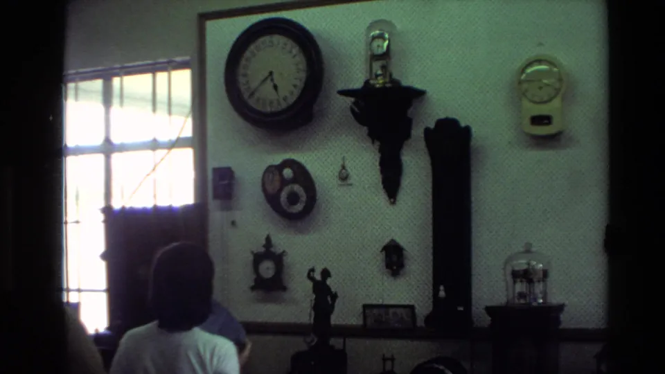 Interior 8mm frame from a private home showing people partly visible below a wall of antique-style clocks and timepieces near a window.