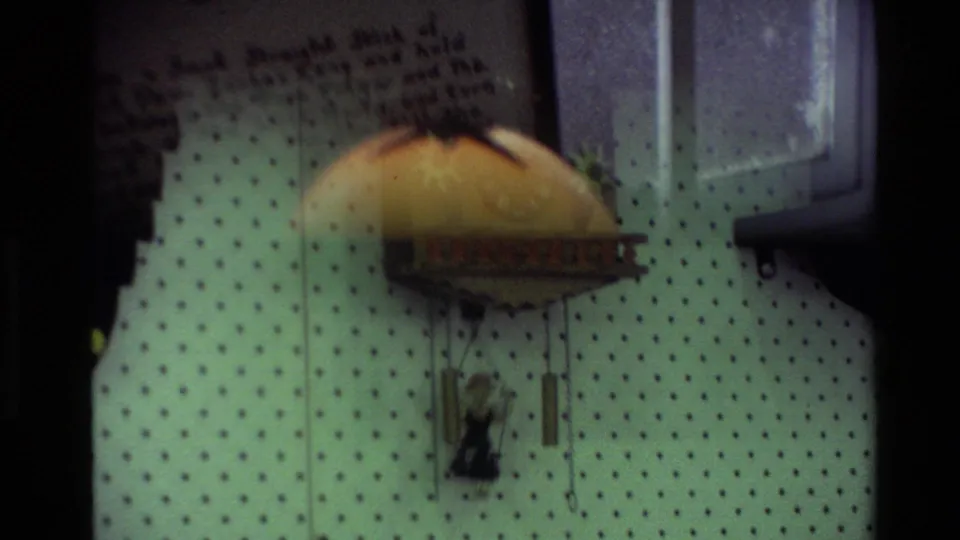 Home-movie close view of patterned wallpaper with a domed hanging decoration and small figure below it in a private New Zealand interior.