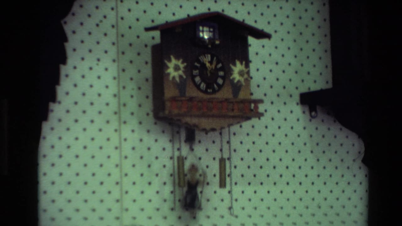 1981 New Zealand home-movie frame showing a cuckoo-style wooden wall clock with star decorations on dotted interior wallpaper.