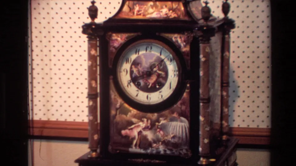 1981 New Zealand home-movie frame showing an ornate wooden clock case with painted figures, columns, and a round clock face.