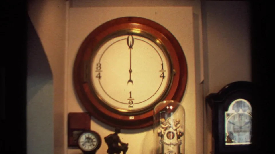 1981 New Zealand 8mm frame focused on a large round wall clock above a clustered display of vintage clocks and glass-covered objects.