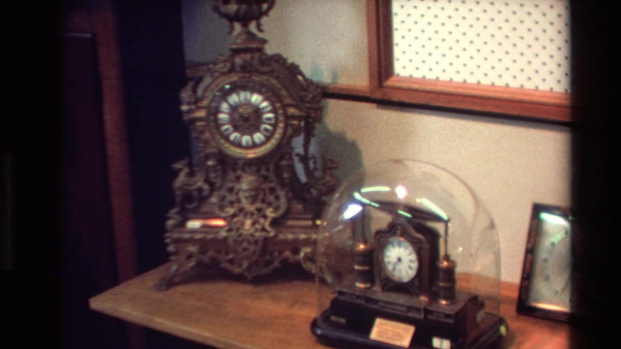 1981 New Zealand home-movie frame showing ornate antique clocks on a wooden shelf beside patterned wall covering.