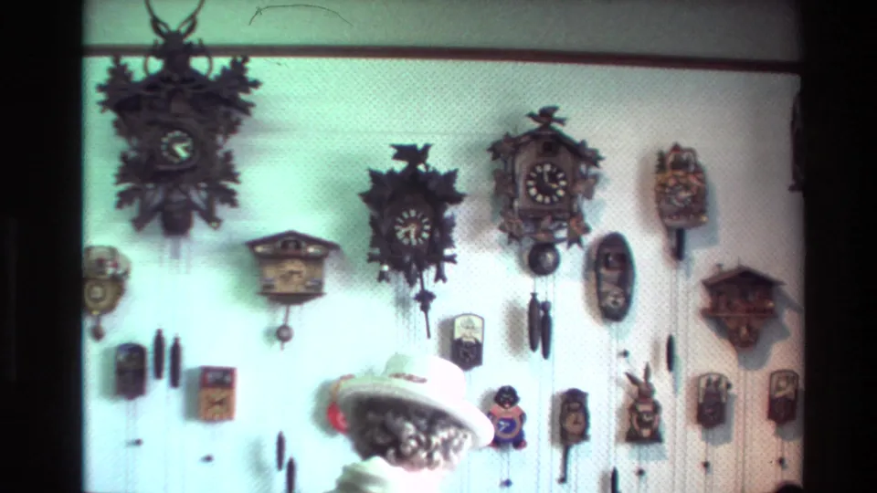 1981 New Zealand home-movie frame showing a wall of cuckoo and hanging clocks with a small hat-wearing figure below.