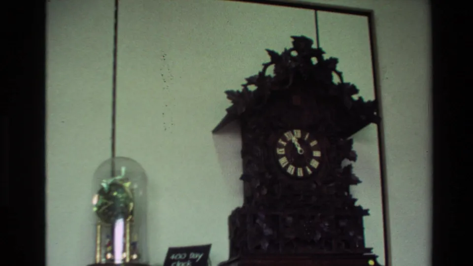 Close view from a 1981 New Zealand 8mm clip of an ornate dark cuckoo clock displayed beside a smaller object under a glass dome.