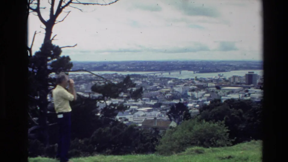 In metadata-supported 1981 New Zealand footage, a man stands on a grassy overlook using binoculars toward a town and broad waterway.