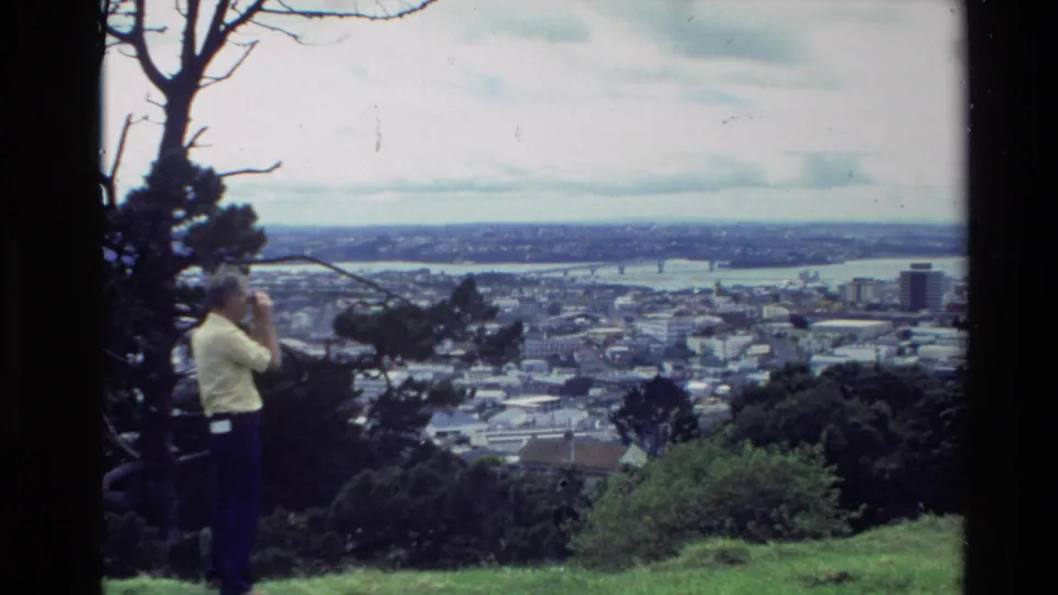 A man remains at a grassy New Zealand overlook with binoculars, framed by trees and a distant town under cloudy sky.