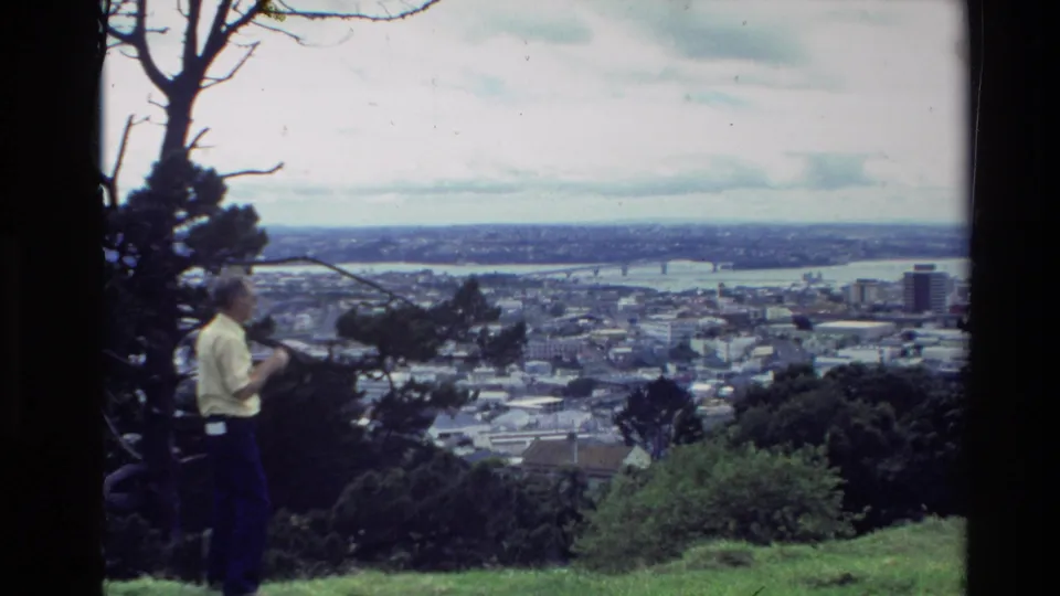 1981 New Zealand home-movie frame showing a man on a hillside viewpoint looking across a city, harbor water, and distant bridge.