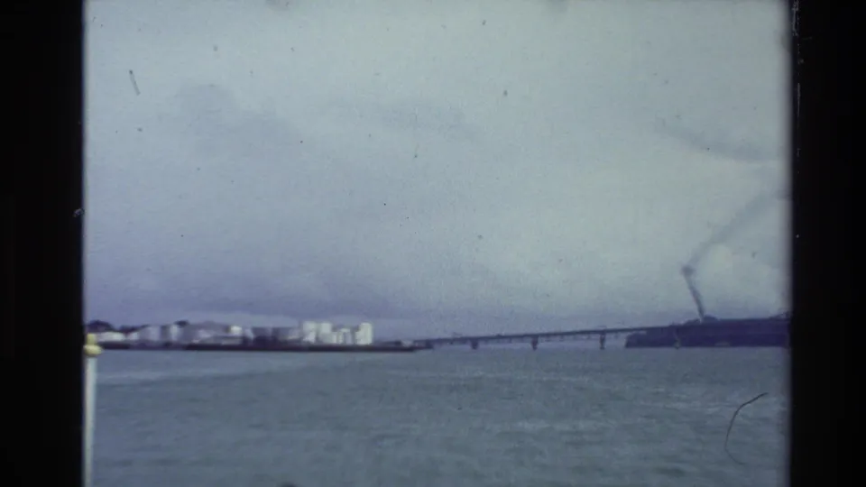 1981 New Zealand home-movie frame looking across harbor water toward shoreline buildings and a long bridge under cloud cover.