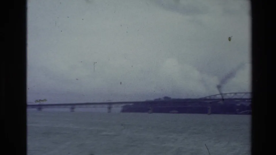 1981 New Zealand harbor view showing open water, a distant bridge span, dark shoreline, and cloudy sky in 8mm film texture.