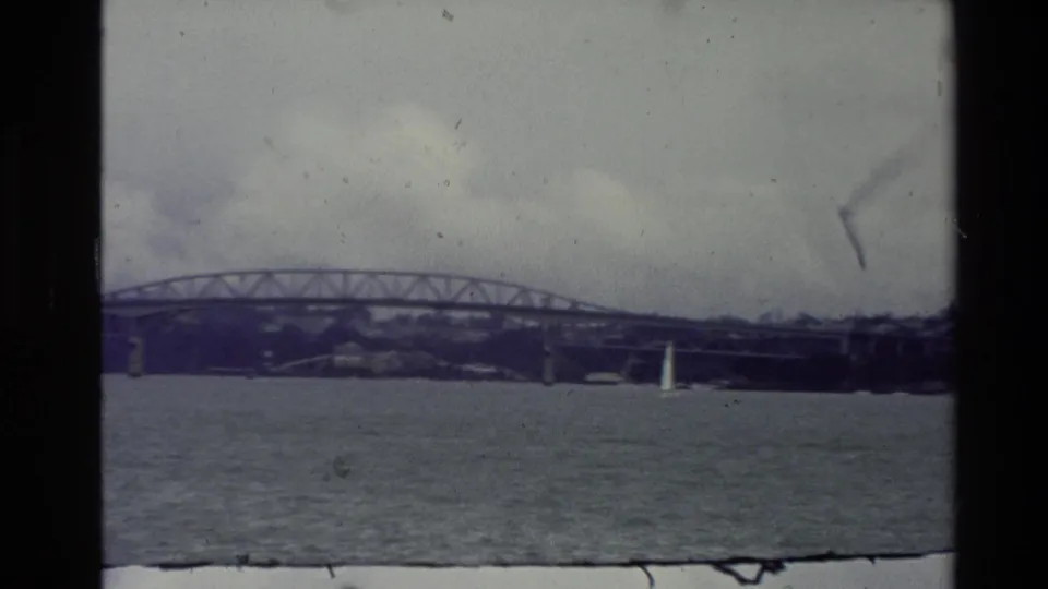 1981 New Zealand home-movie frame with a sailboat on rippled water beneath a long steel truss bridge.