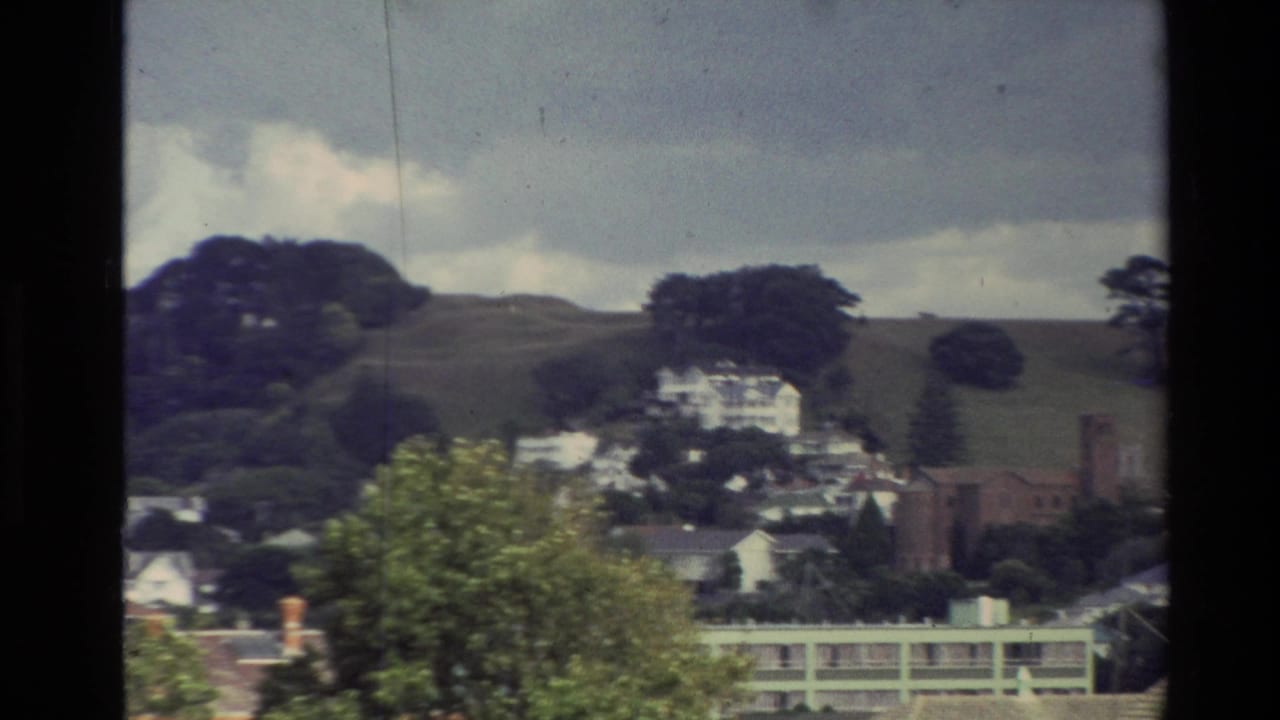 1981 New Zealand home-movie view through a window toward hillside houses, trees, and a cloudy sky.