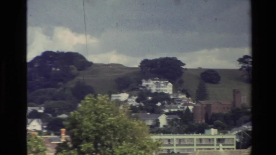1981 New Zealand home-movie view through a window toward hillside houses, trees, and a cloudy sky.